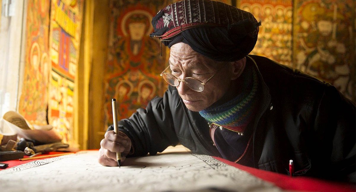 A man dressed as an ancient scribe wearing glasses, writing with a quill pen on parchment.
