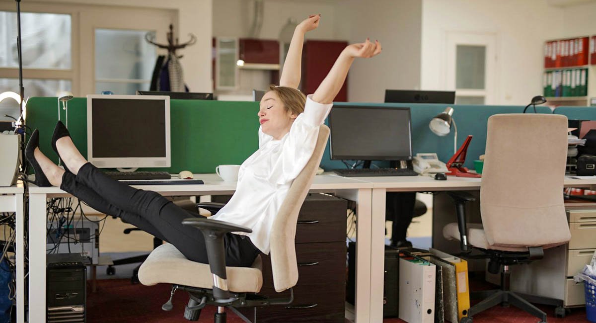 I did it! White woman in office stretching back in her chair with arms over her head