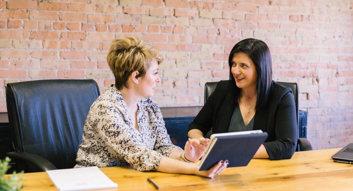 What's your answer to this question? Two businesswomen talking at a desk with a tablet in hand