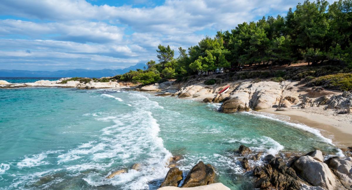 Aegean sea coast with greenery around, rocks, bushes and trees, blue water with waves, Greece