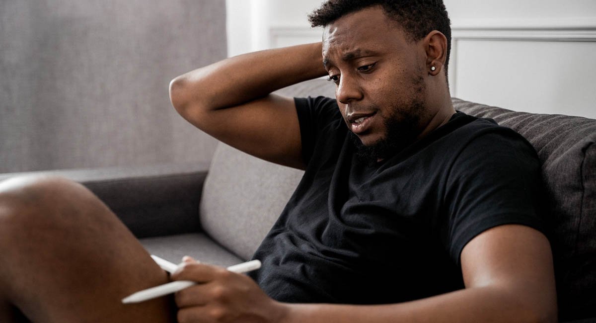 Man in black t-shirt sitting on couch writing looking confused